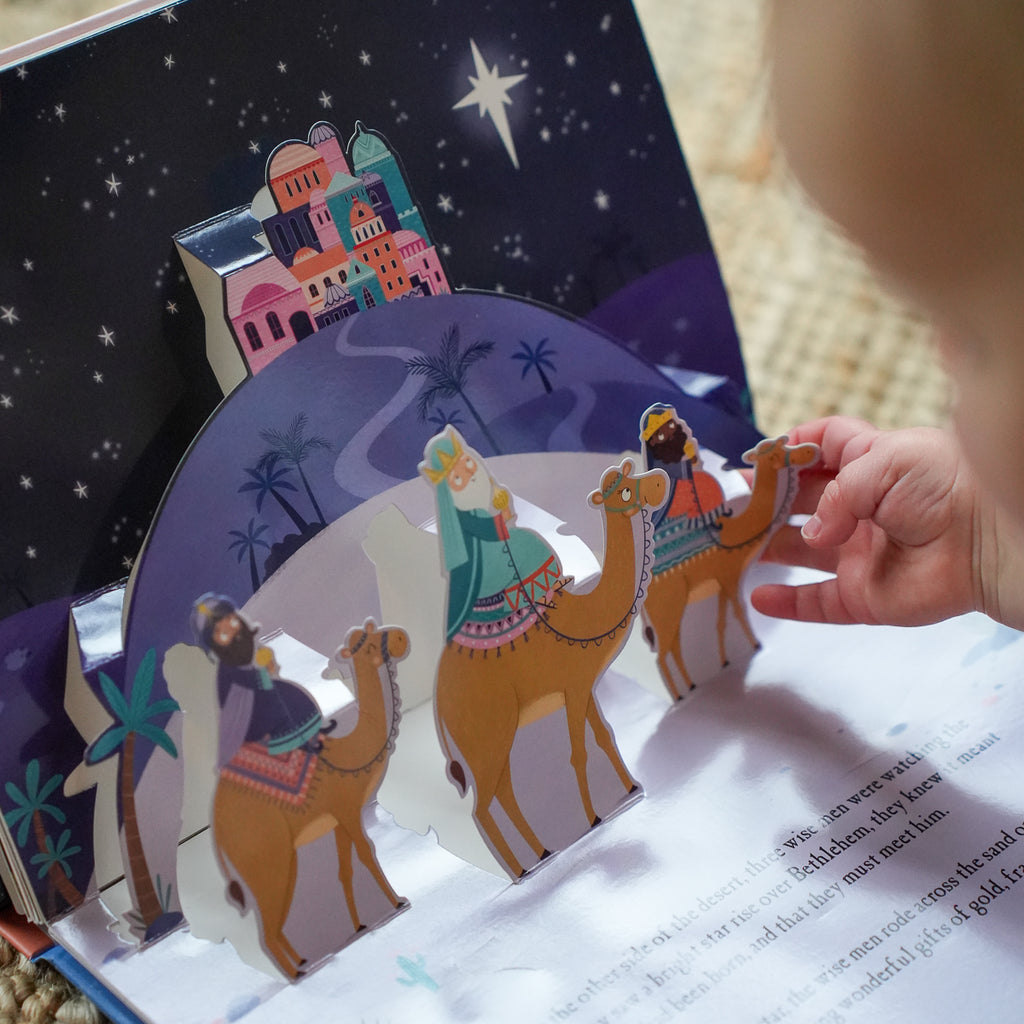 A close-up of a child's hand opening a 3D pop-up children's book about the Holy Night, showing illustrated camels, figures, and a starry night sky background.