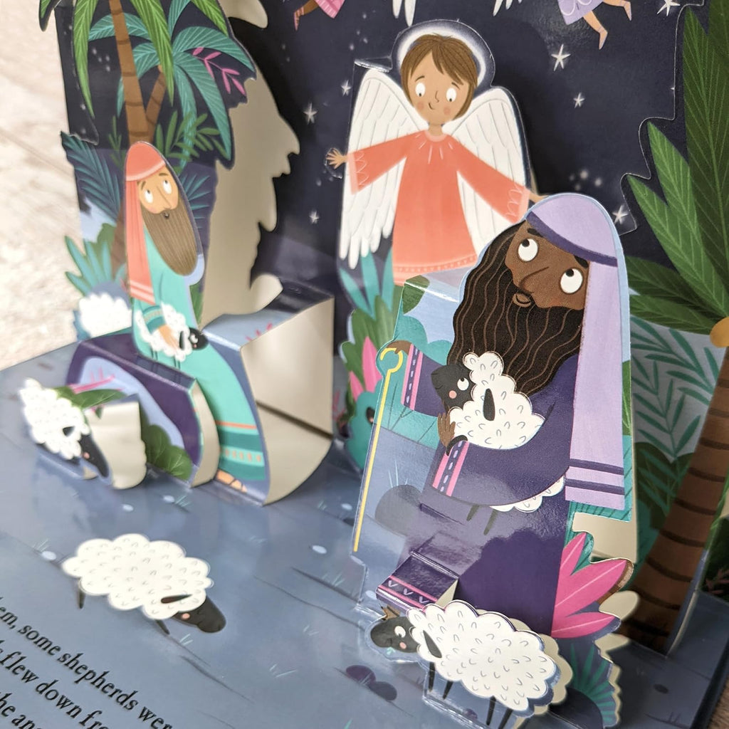 A colorful 3D pop-up cardboard Christmas nativity scene featuring illustrated figures, palm trees, clouds, and a dark starry backdrop.