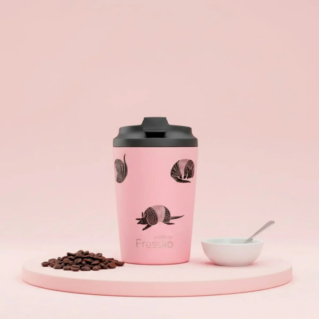Pink coffee tumbler with black lid on a pink platform with coffee beans and a bowl.