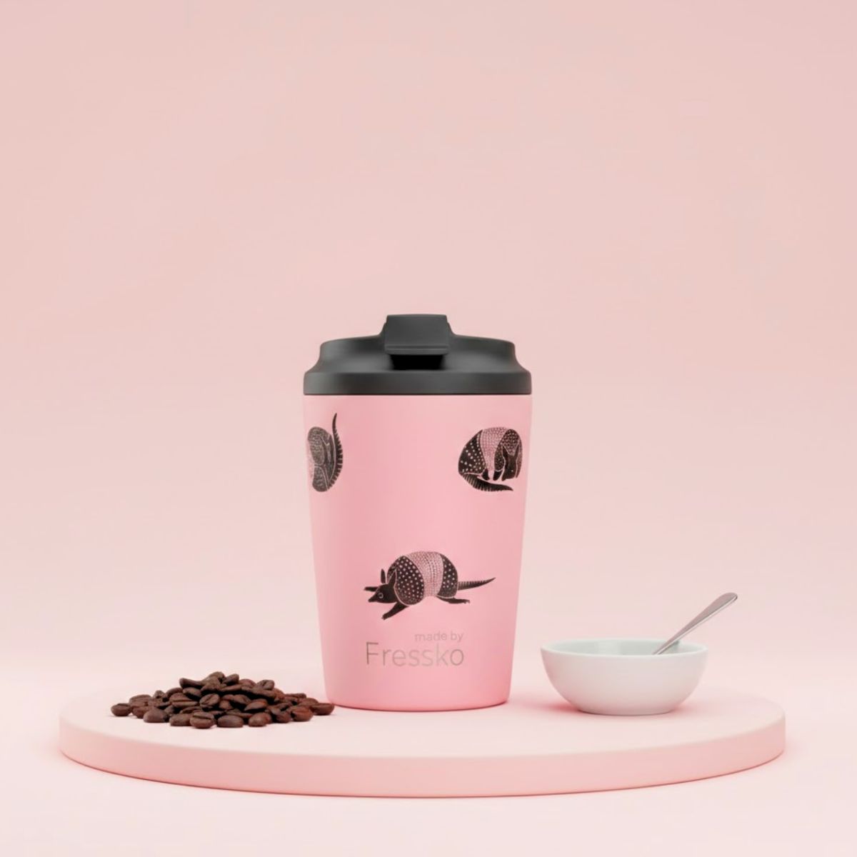 Pink coffee tumbler with black lid on a pink platform with coffee beans and a bowl.