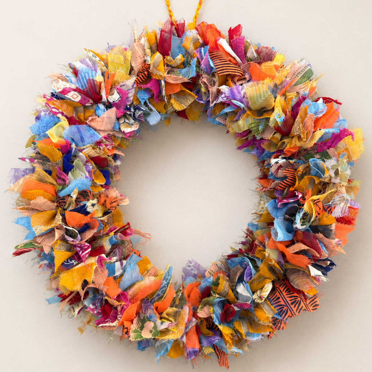 A handmade fabric wreath made from many colorful cut-up textile pieces, hanging against a plain light beige wall.