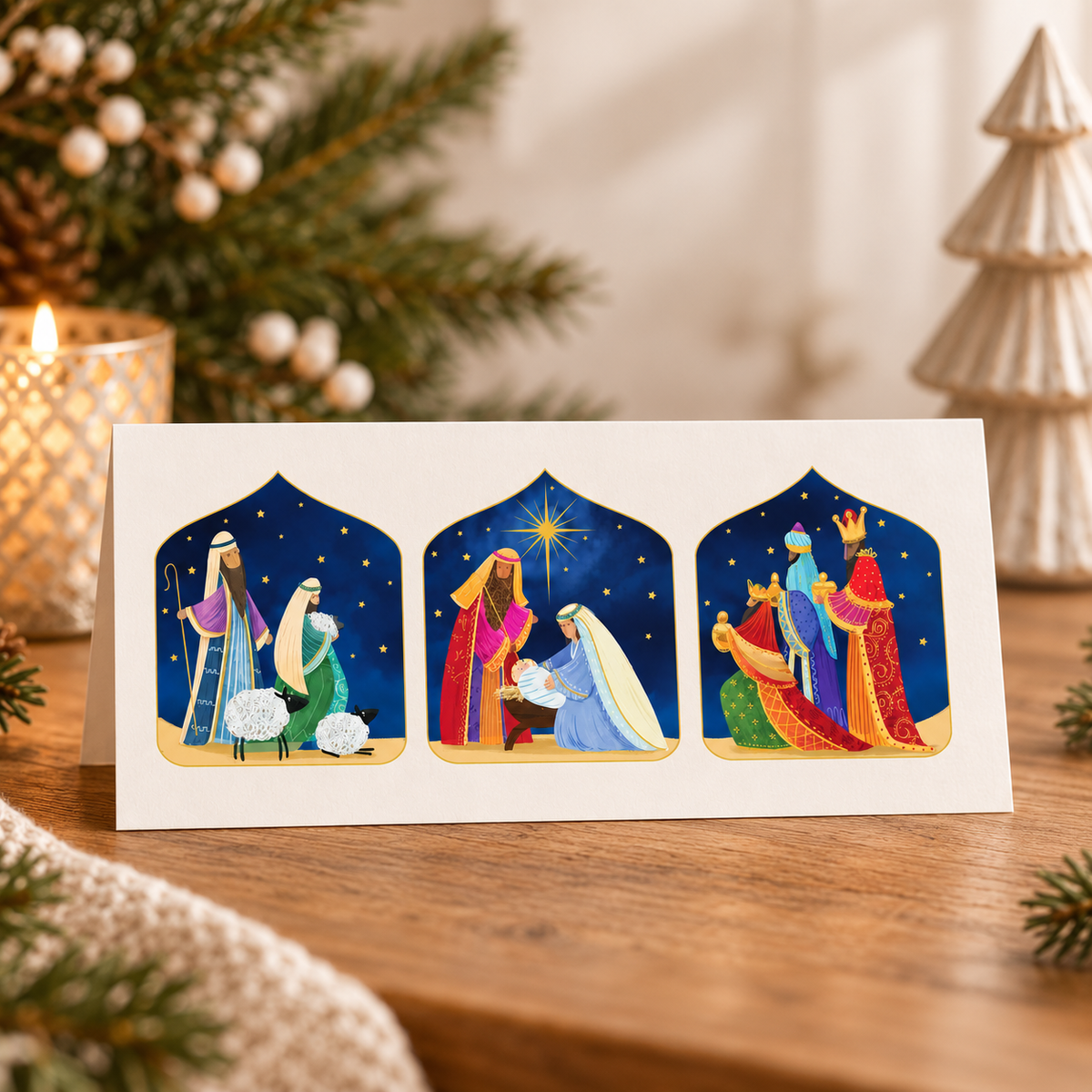 A white greeting card with three colorful Christmas nativity scenes, displayed on a wooden table with holiday decorations in the background.