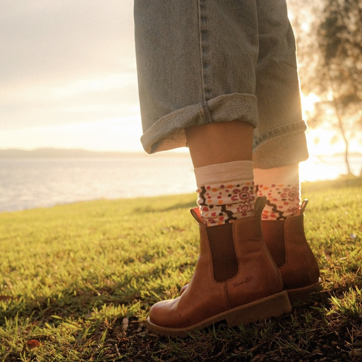 A person standing outdoors on grass near water at sunset, wearing light blue cuffed jeans, patterned socks, and brown Chelsea-style boots.