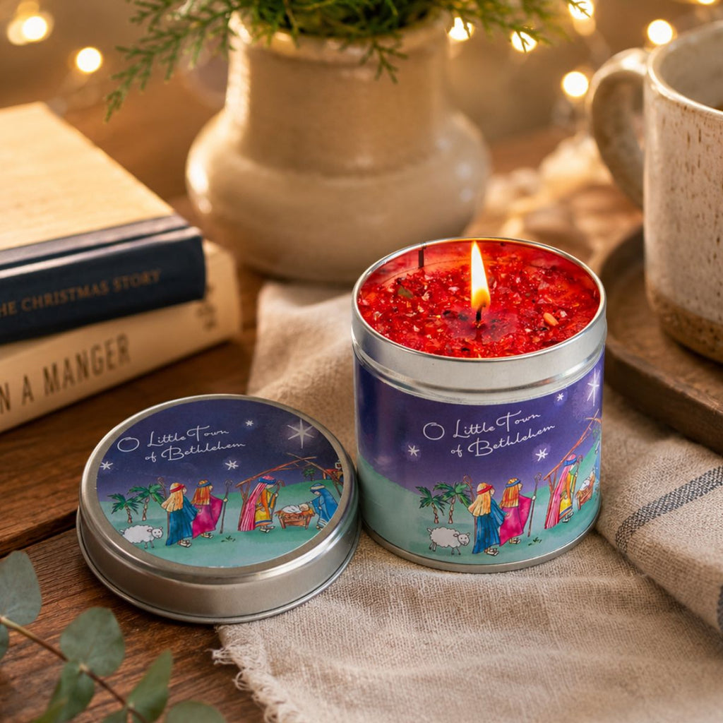A lit red wax candle in a silver tin with a matching lid placed beside it on a fabric surface. The lid and candle label read "Little Star of Bethlehem" and show a nativity scene.