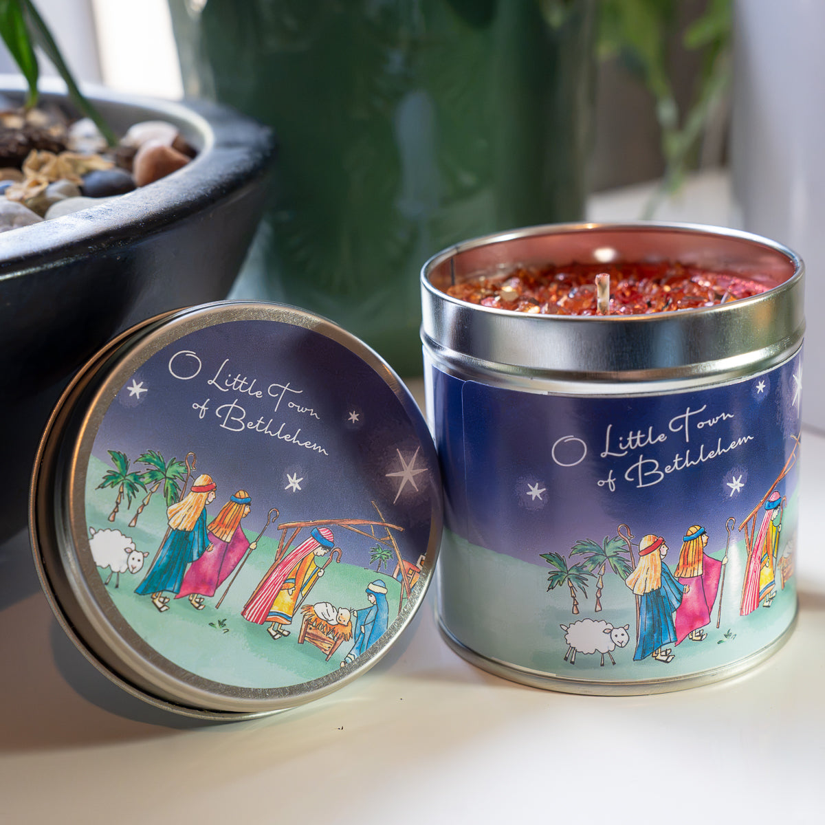 A decorative metal candle tin with its lid leaning beside it on a tabletop. The open tin shows red wax and a blue illustrated label reading 'Little Town of Bethlehem,' while the lid has a matching pastel nativity scene.