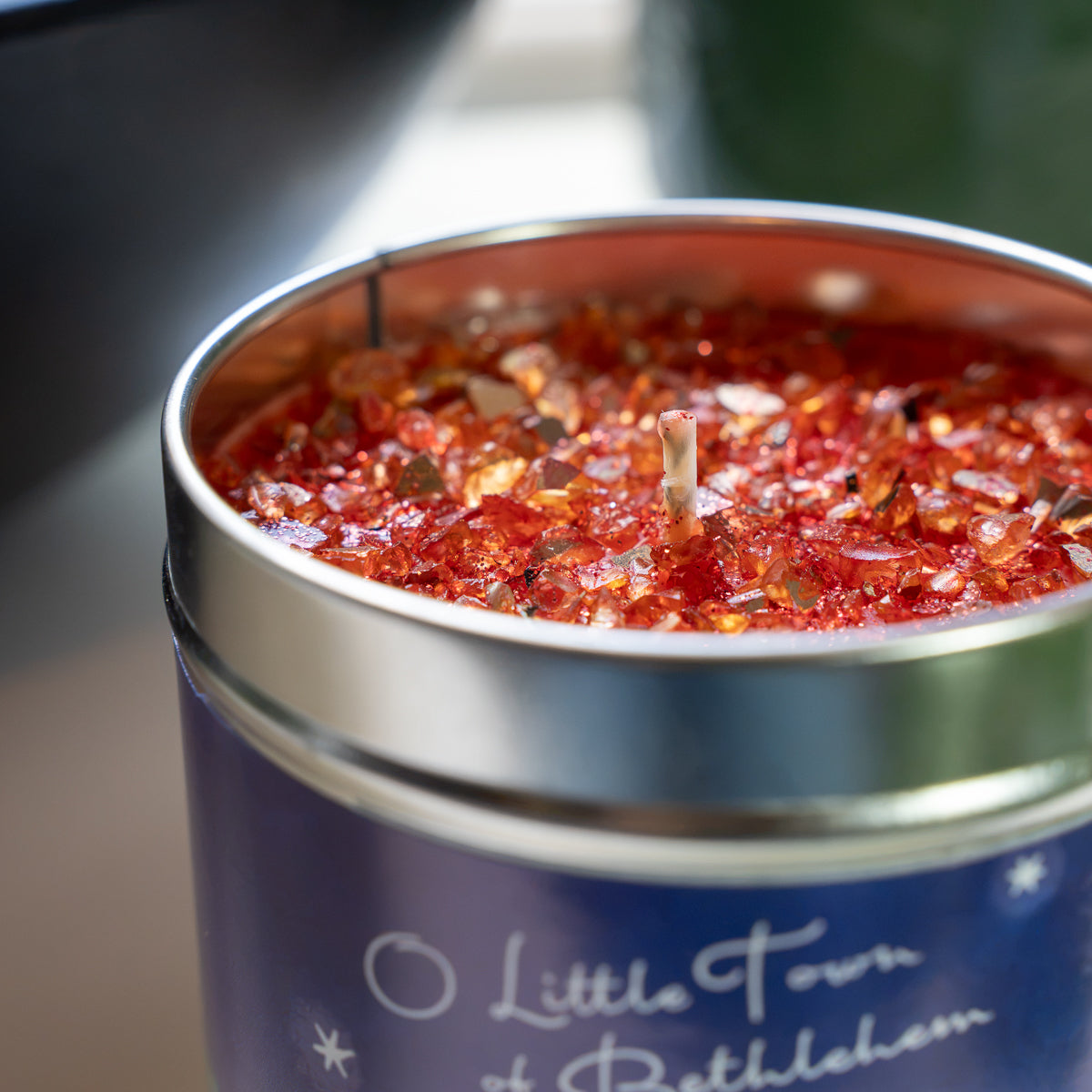 A close-up of a tin candle with a navy blue label reading 'Little Town of Bethlehem' and a silver metal container filled with red wax and glittery flakes.