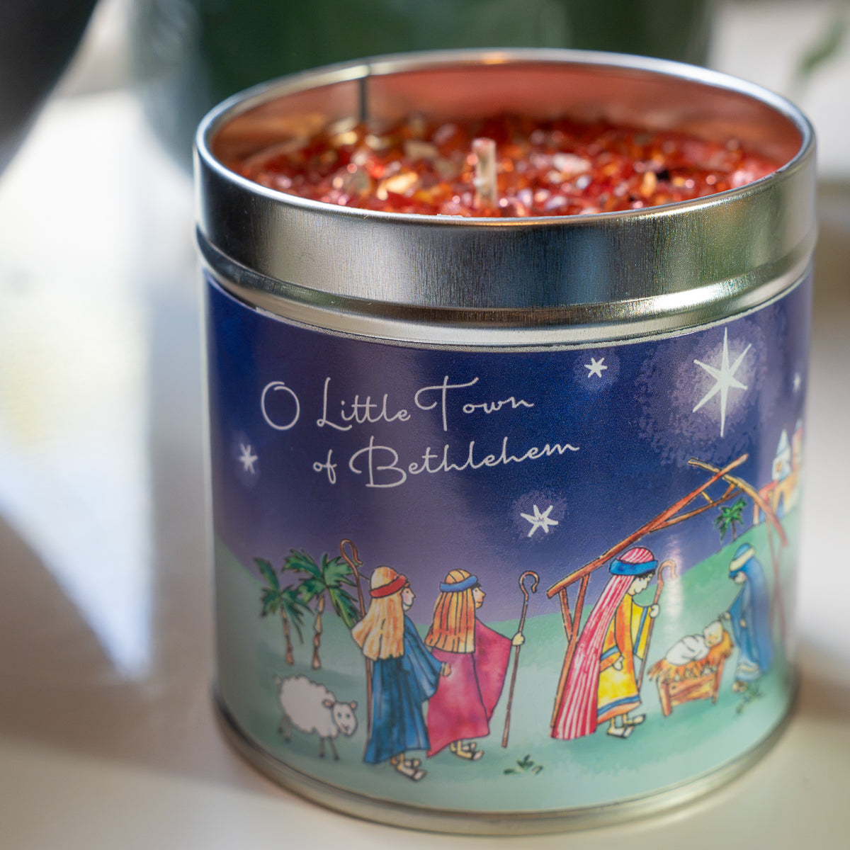 A tin candle with a silver metal lid and a colorful printed label reading 'O Little Town of Bethlehem,' filled with red wax and wicks, photographed on a light surface with a softly blurred background.