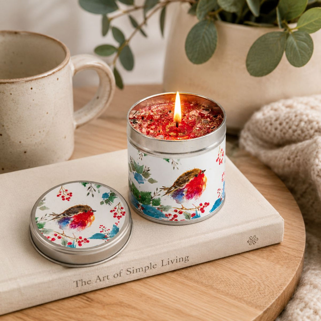 A lit metal tin candle with a colorful bird illustration sits on a book on a wooden surface, with a matching lid beside it. A ceramic mug, plant, and knitted textile are softly blurred in the background.