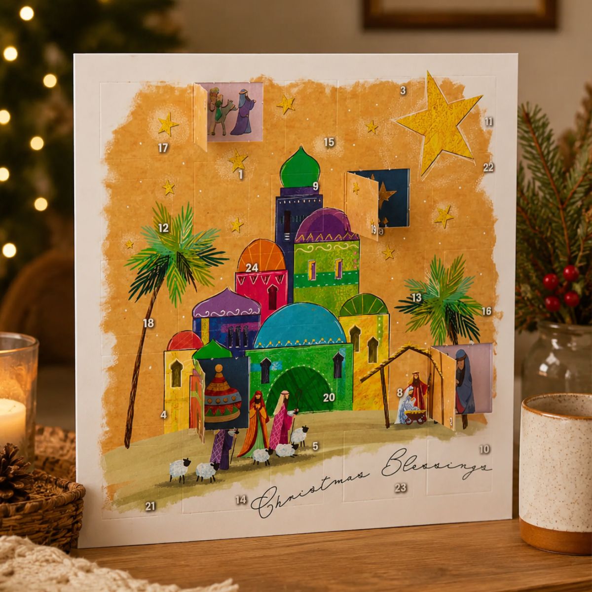 A Christmas Blessing advent calendar card displayed on a wooden surface with warm holiday decor in the background. The card features a colorful nativity scene with numbered doors, a large gold star, palm trees, and the text 'Christmas Blessing' at the bottom.
