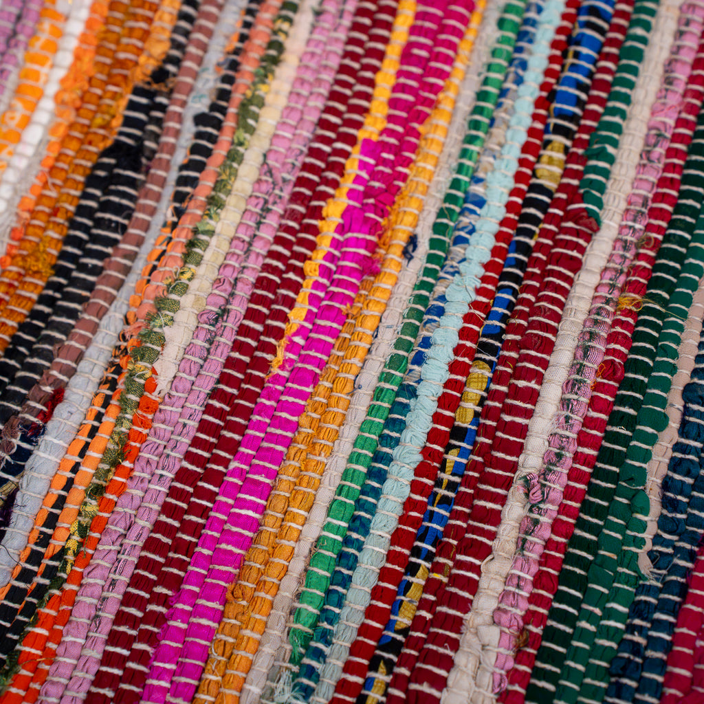 A close-up of a woven multicolor rug or textile with diagonal stripes in orange, black, white, pink, yellow, green, blue, red, and dark teal.