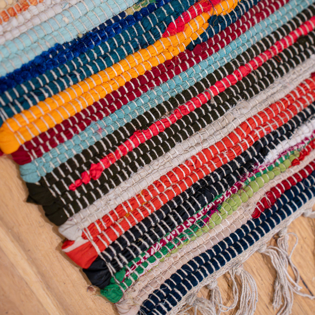 A close-up of a handmade woven rag rug with colorful horizontal stripes in blue, yellow, red, green, black, and beige, laid out on a light wood floor.