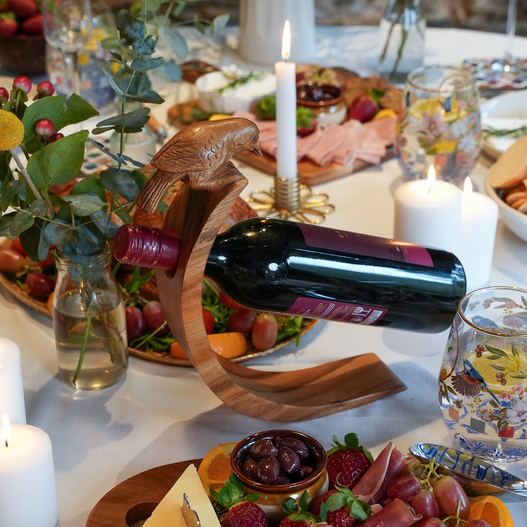 A dark red wine bottle rests horizontally in a carved wooden wine bottle holder shaped like a bird perched on a branch, set on a decorated dining table with candles, food, and glassware.