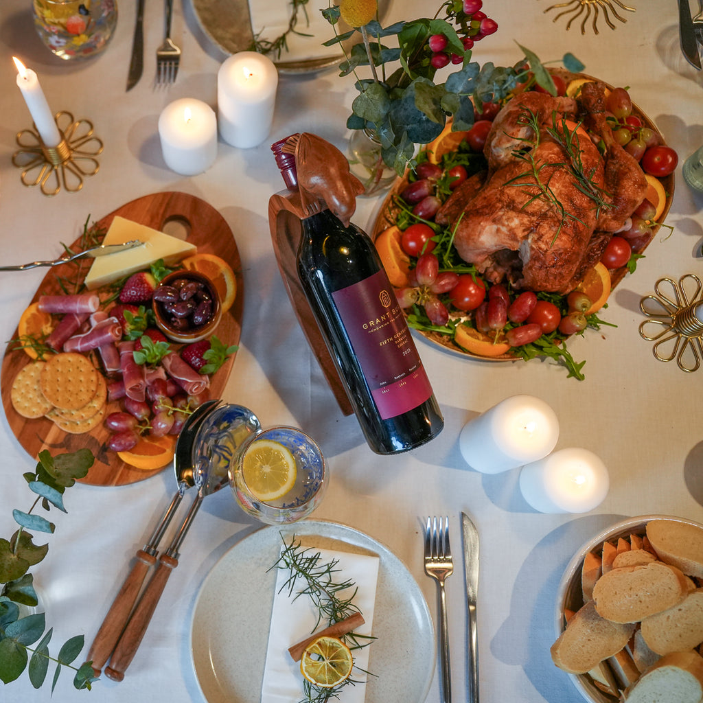 A styled holiday dining table setting featuring a roasted turkey on an orange and grapefruit garnish platter, sliced cured meats, crackers, bread, candles, and a bottle of red wine on a white tablecloth.