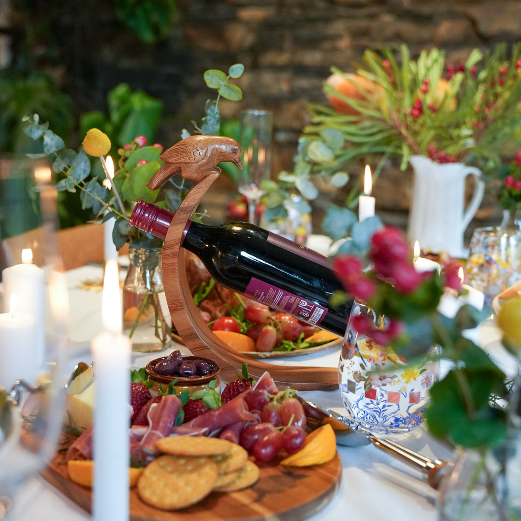A dark red wine bottle displayed in a wooden bird-shaped holder on a decorated dining table with candles, flowers, crackers, and charcuterie items.