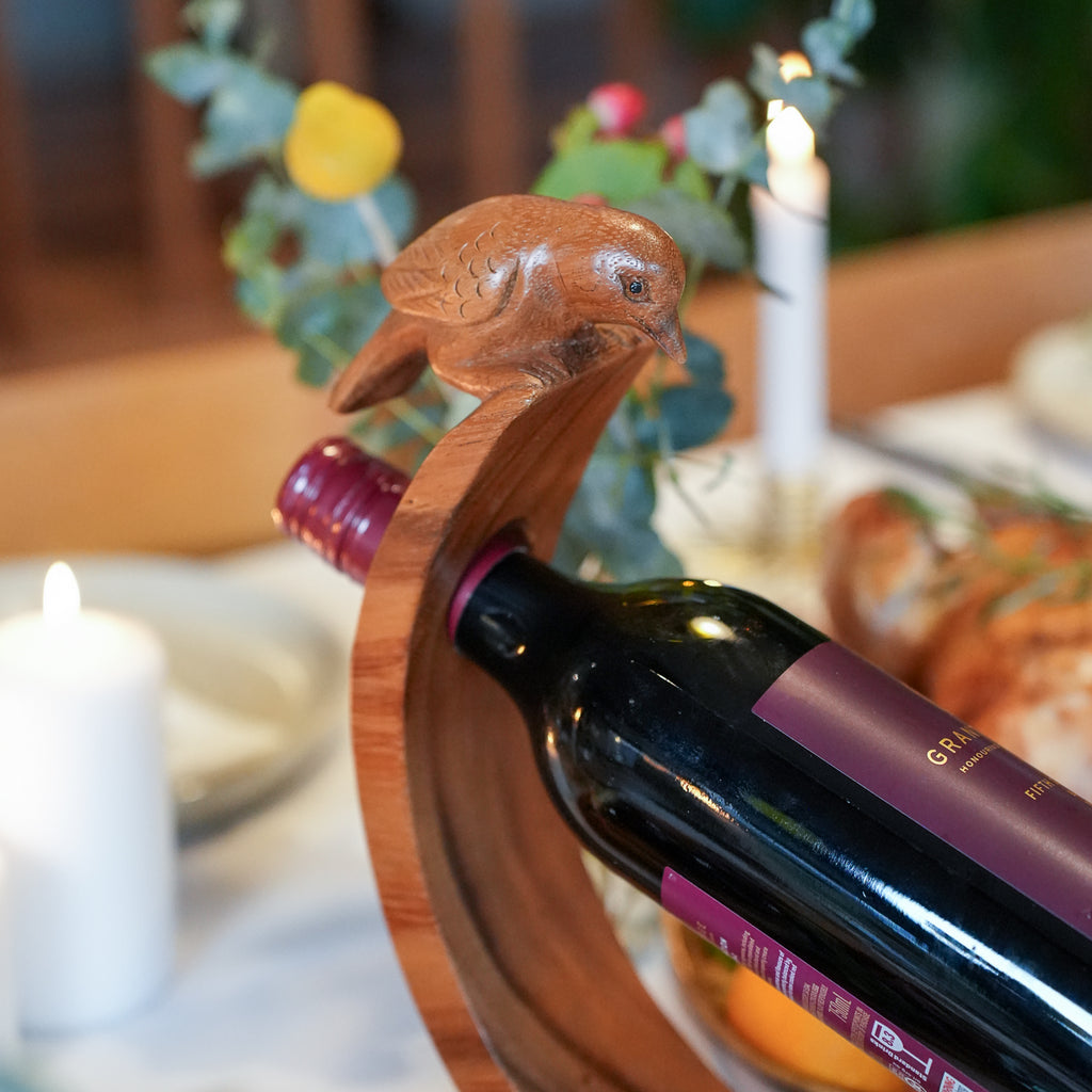 A close-up of a wine bottle resting in a curved wooden holder shaped like a bird, set on a softly blurred dining table with lit candles and flowers.