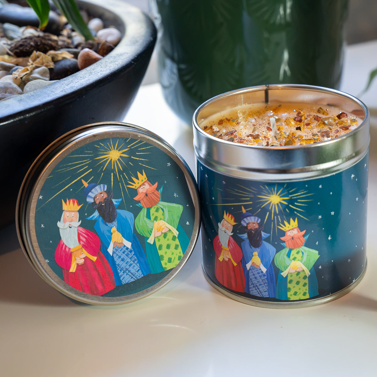 A decorative metal candle tin with its lid placed beside it on a tabletop. The tin is open and shows a scented candle with visible wick and wax.