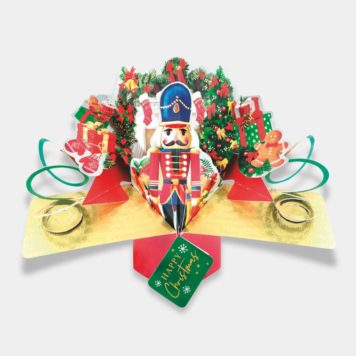 A 3D pop-up cardboard Christmas gift box featuring a nutcracker soldier, a small decorated tree, wrapped presents, gingerbread cookies, and a green hanging tag that reads 'Merry Christmas' on a light gray background.