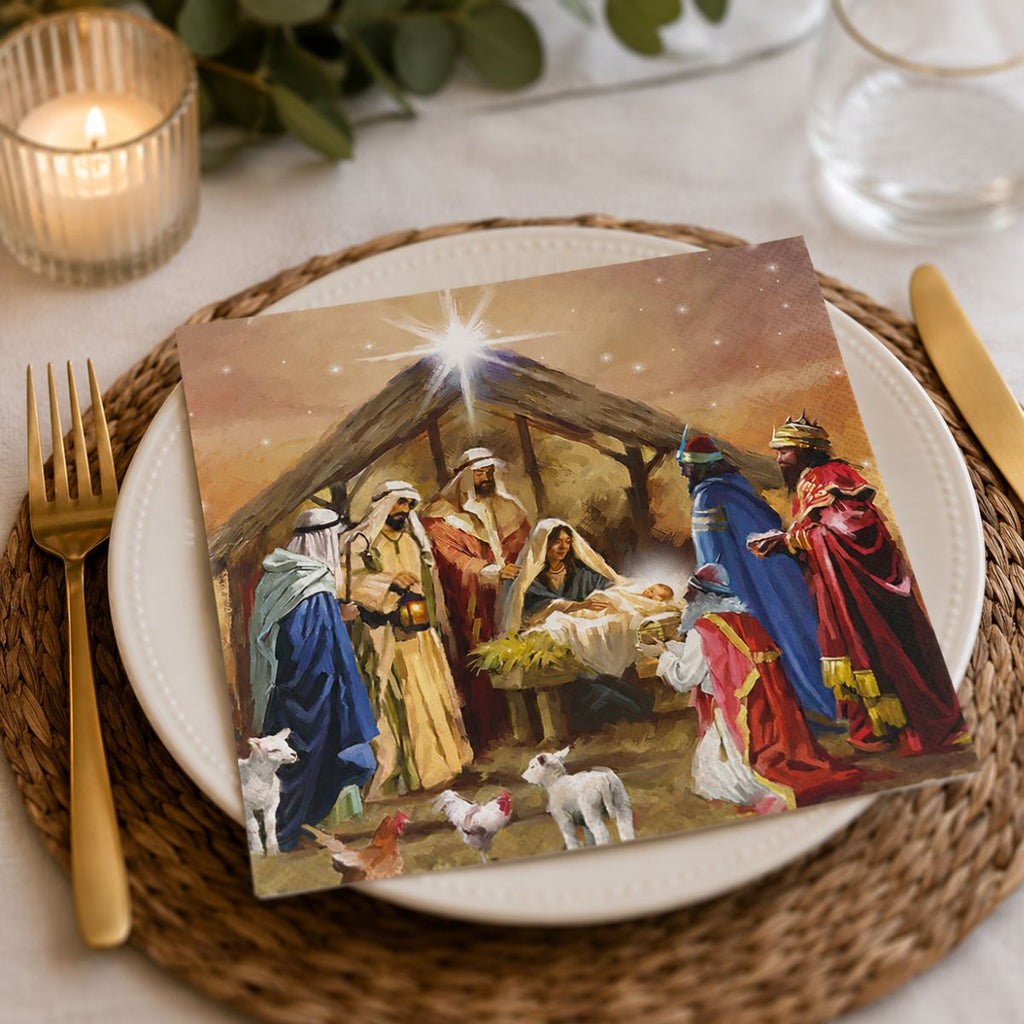 A printed napkin featuring a Nativity scene, styled on a white plate over a woven placemat with gold-toned cutlery and a lit candle in the background.