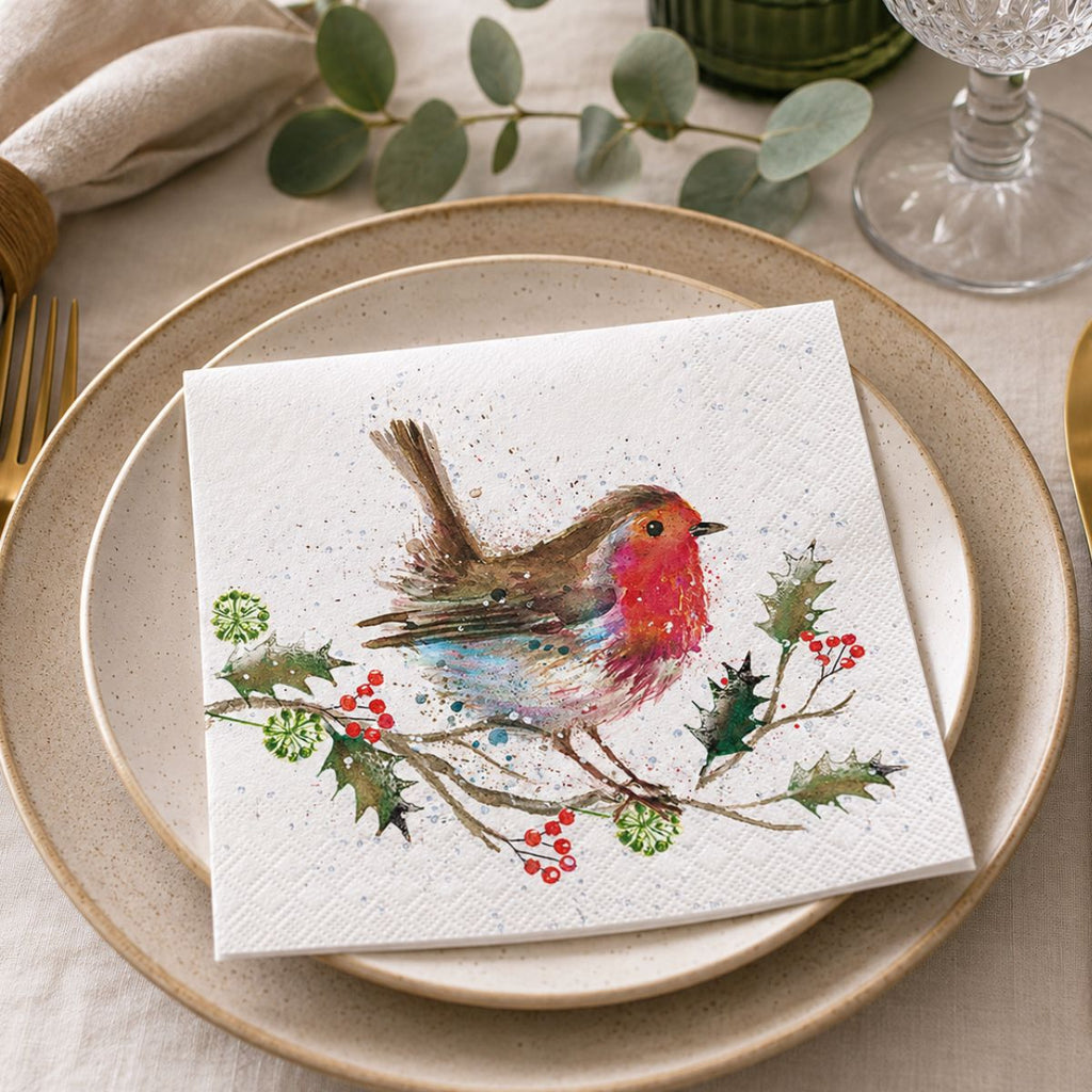 A square white paper napkin featuring a colorful watercolor robin perched on a branch with holly leaves and red berries, styled on top of beige plates at a table setting.