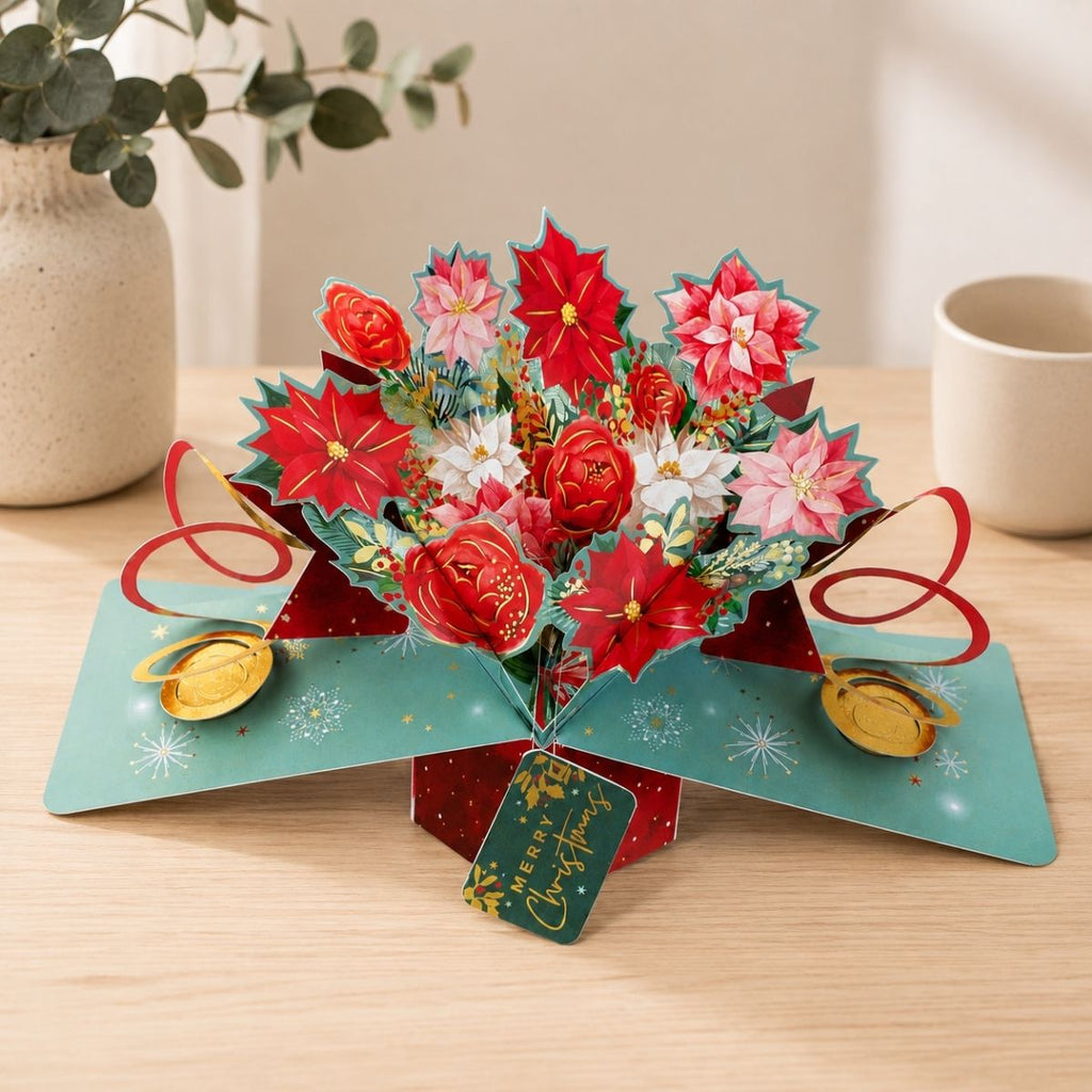 A 3D pop-up cardboard Christmas flower bouquet sits open on a light wood tabletop. The popper features red and pink poinsettias, white flowers, green foliage, red ribbon loops, gold coins, and a green tag that reads 'Merry Christmas.