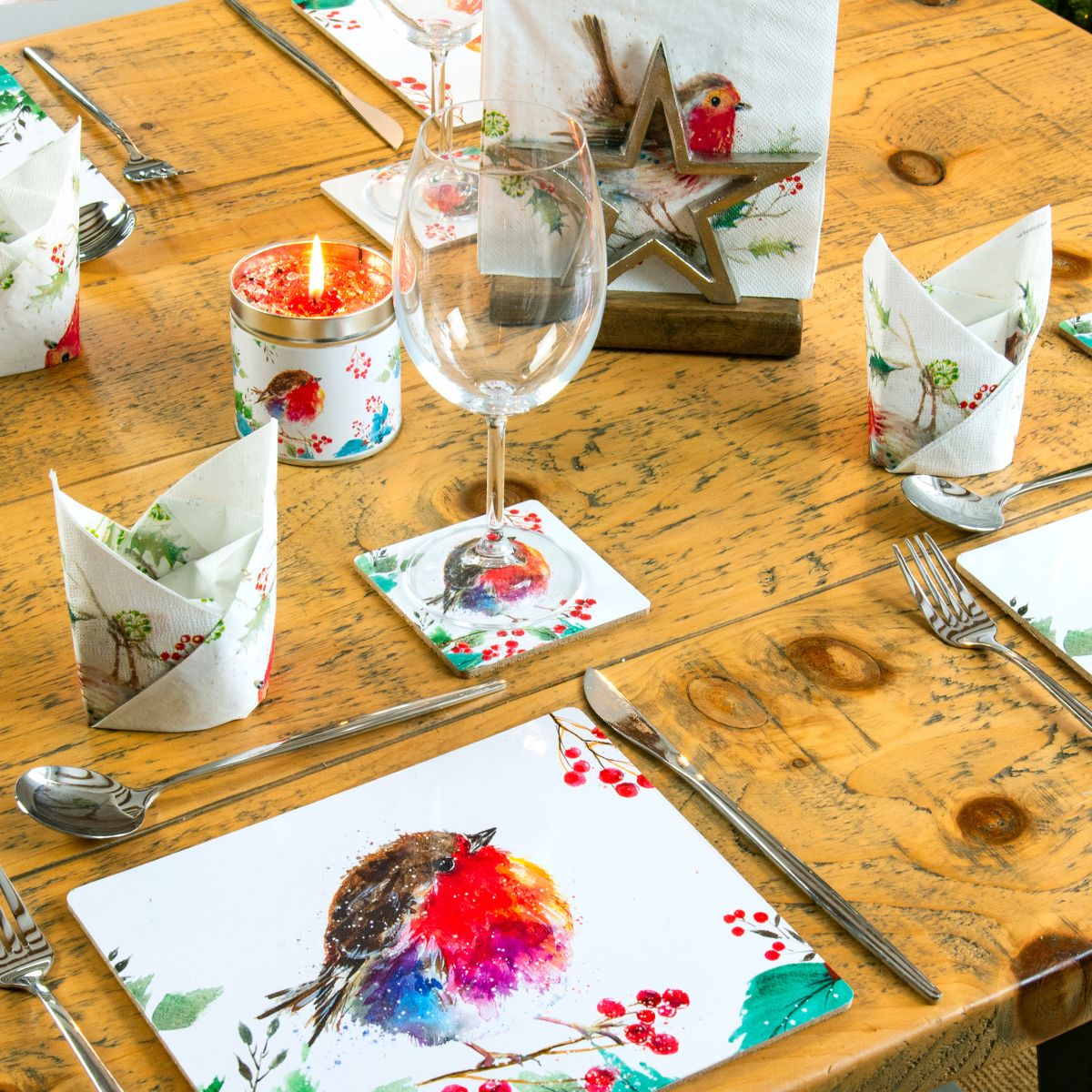 A styled holiday table setting featuring square white placemats printed with colorful bird and berry illustrations, folded napkins, silver cutlery, a wine glass, and a candle on a rustic wooden table.