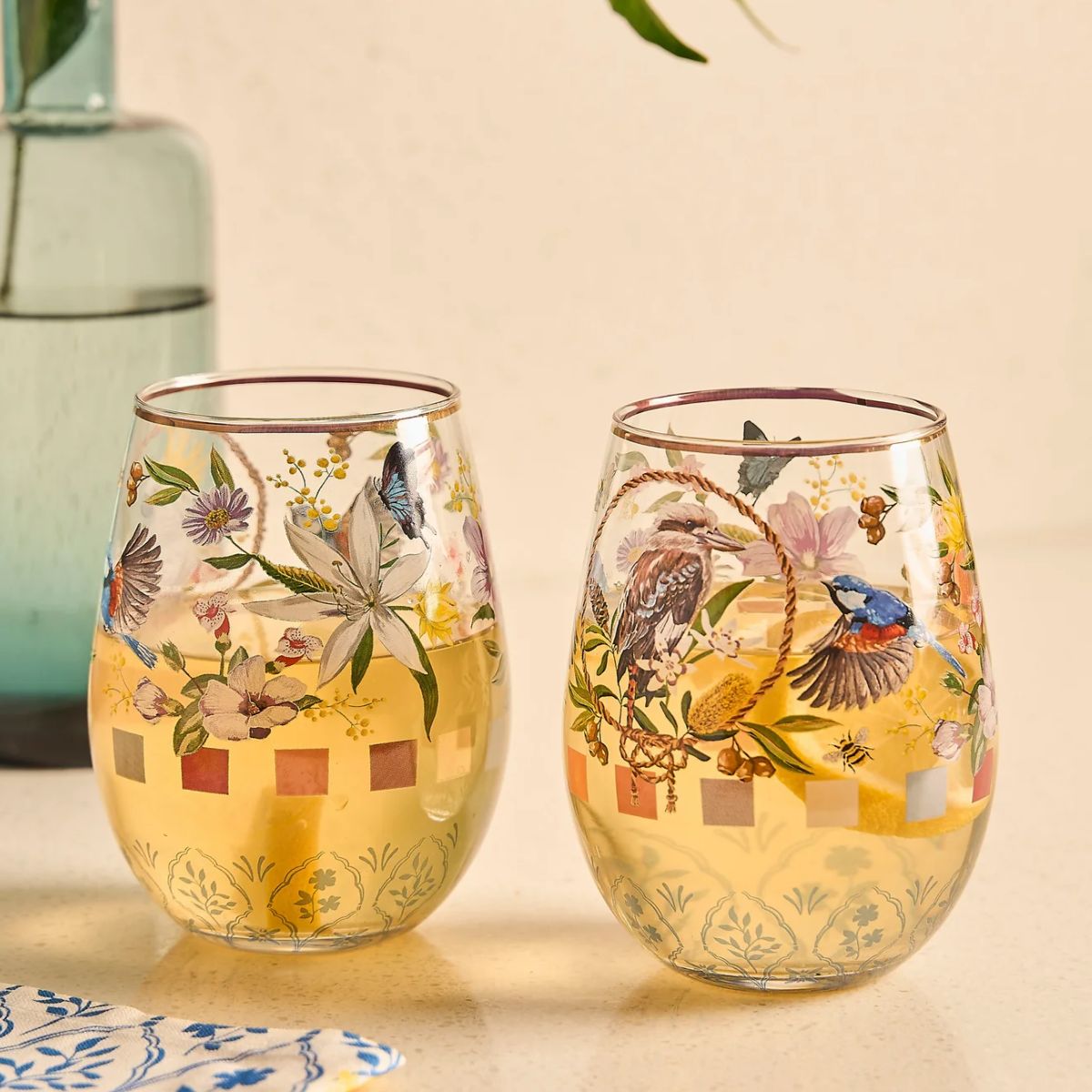 Two stemless wine glasses with floral and bird designs on a beige background