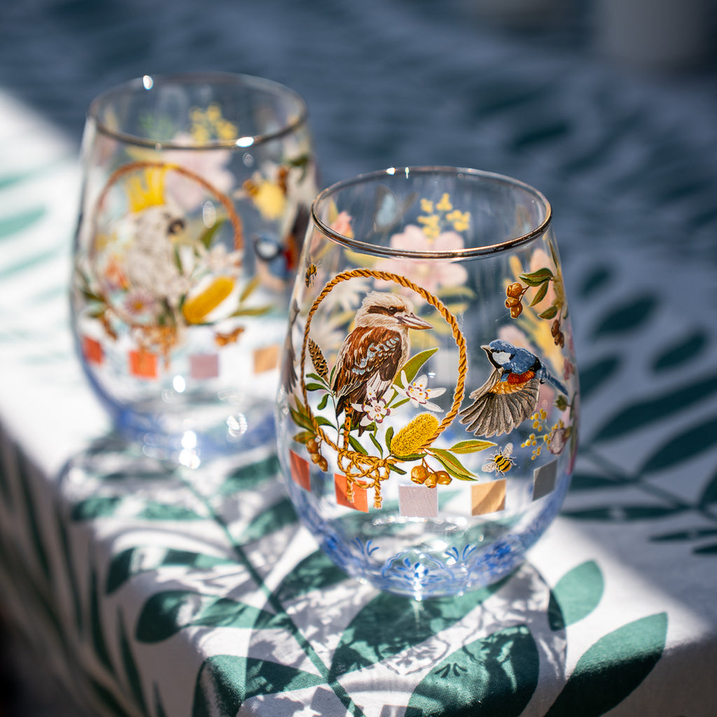 Two clear stemless wine glasses decorated with colorful bird and floral illustrations, set on a white tablecloth with green leaf prints in bright natural light.