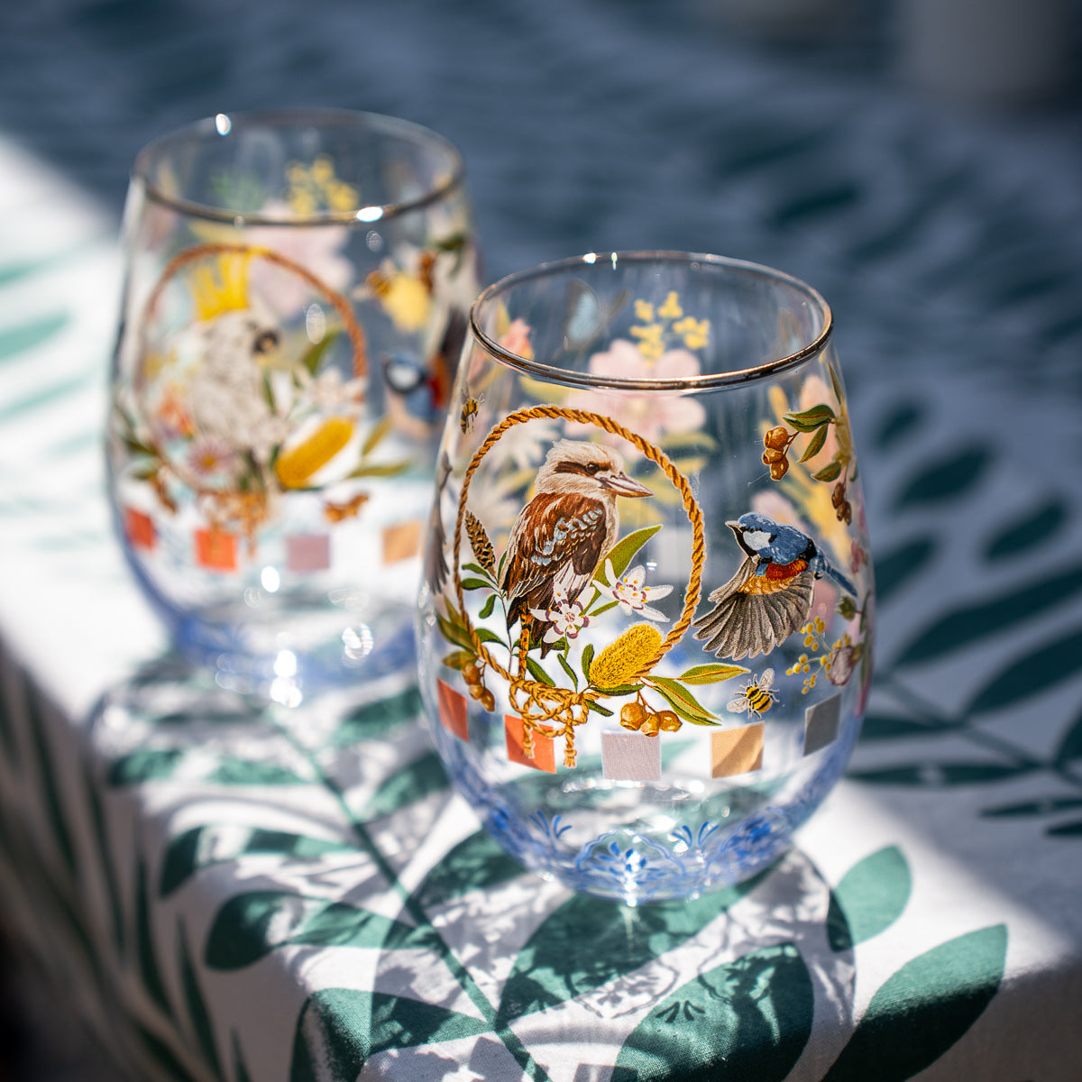 Two clear stemless wine glasses decorated with colorful bird and floral illustrations, set on a white tablecloth with green leaf prints in bright natural light.
