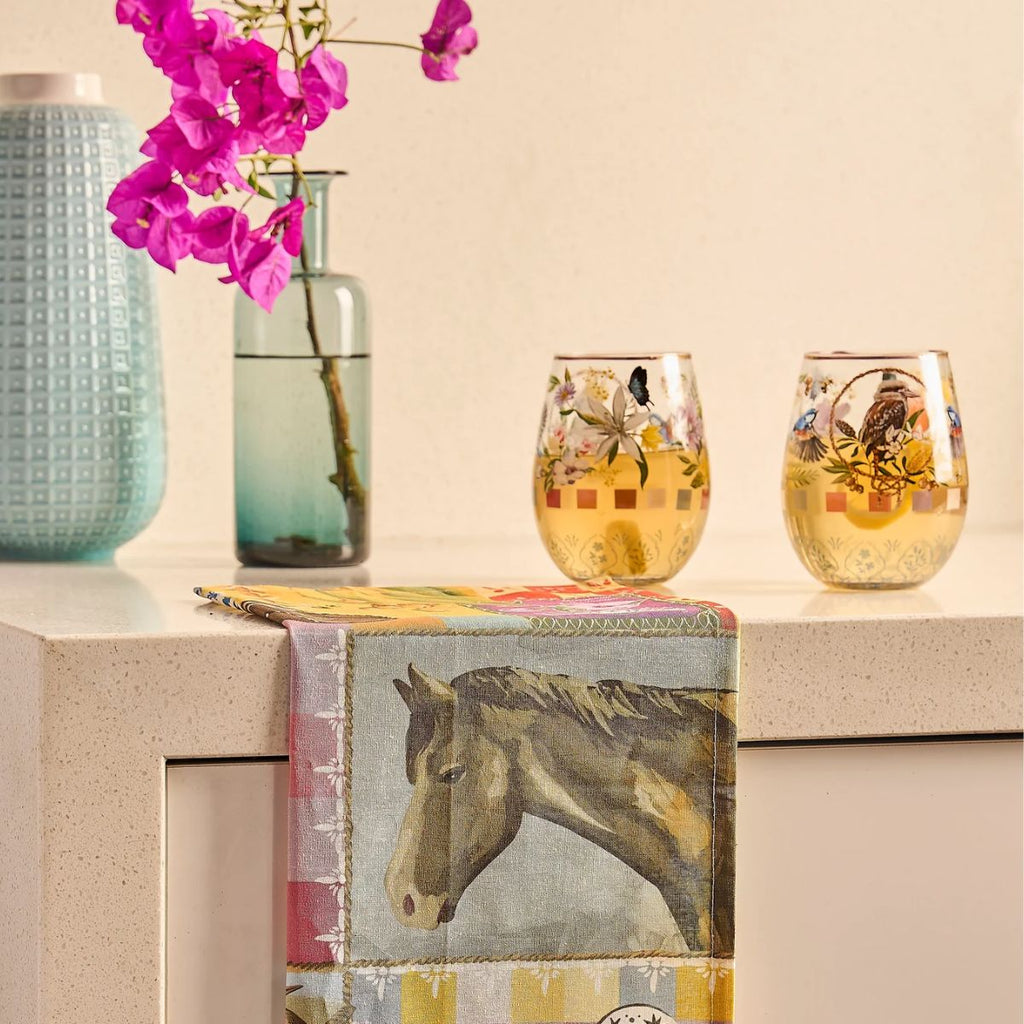 Two floral-patterned stemless glasses on a surface with a horse-themed towel and pink flowers in the background.