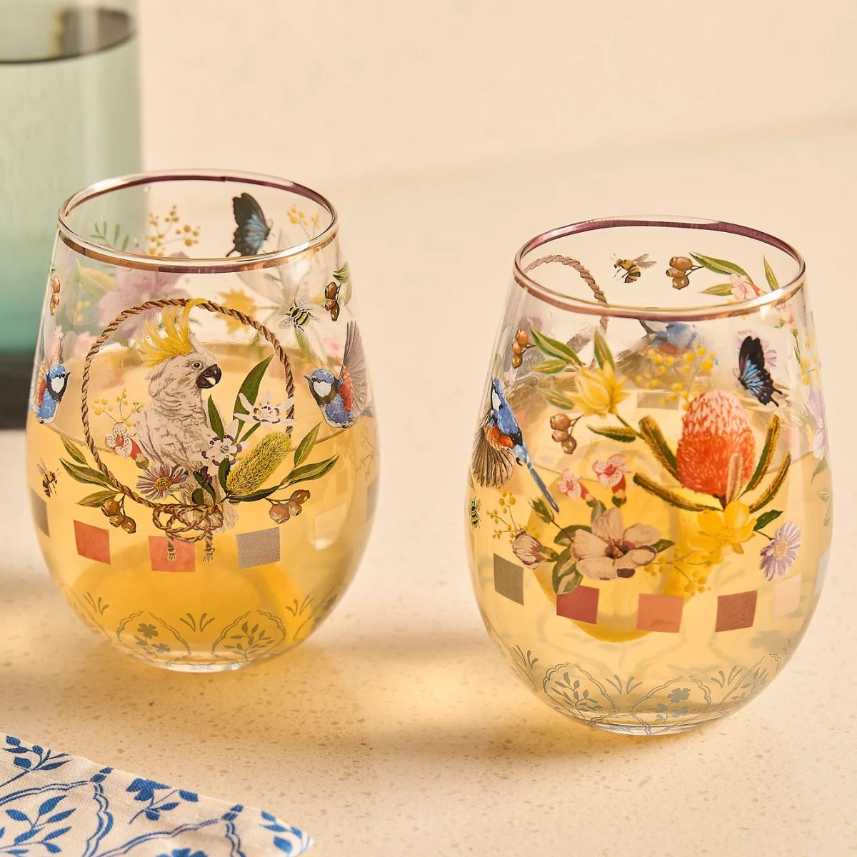 Two stemless wine glasses with decorative parrot and floral patterns on a beige background.