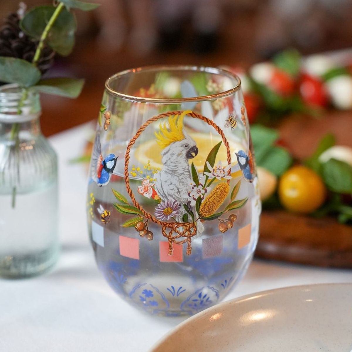 A clear stemless wine glass decorated with a colorful parrot, flowers, bees, and tropical motifs, set on a white table with a blurred dining background.
