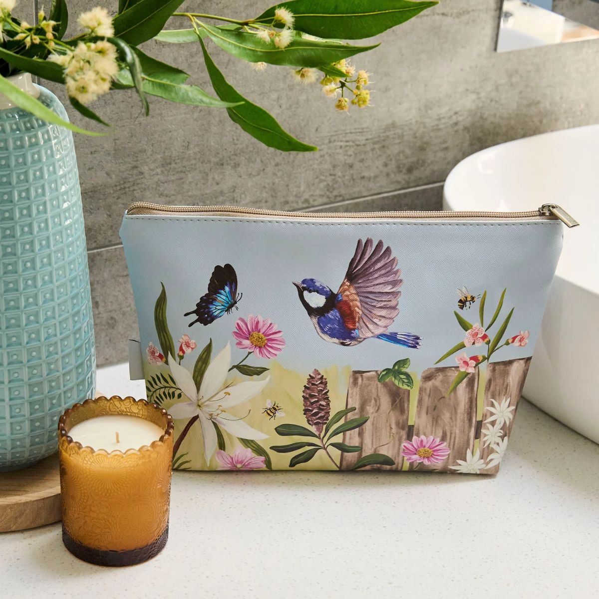 Cosmetic bag with nature-themed design next to a candle and vase on a surface.