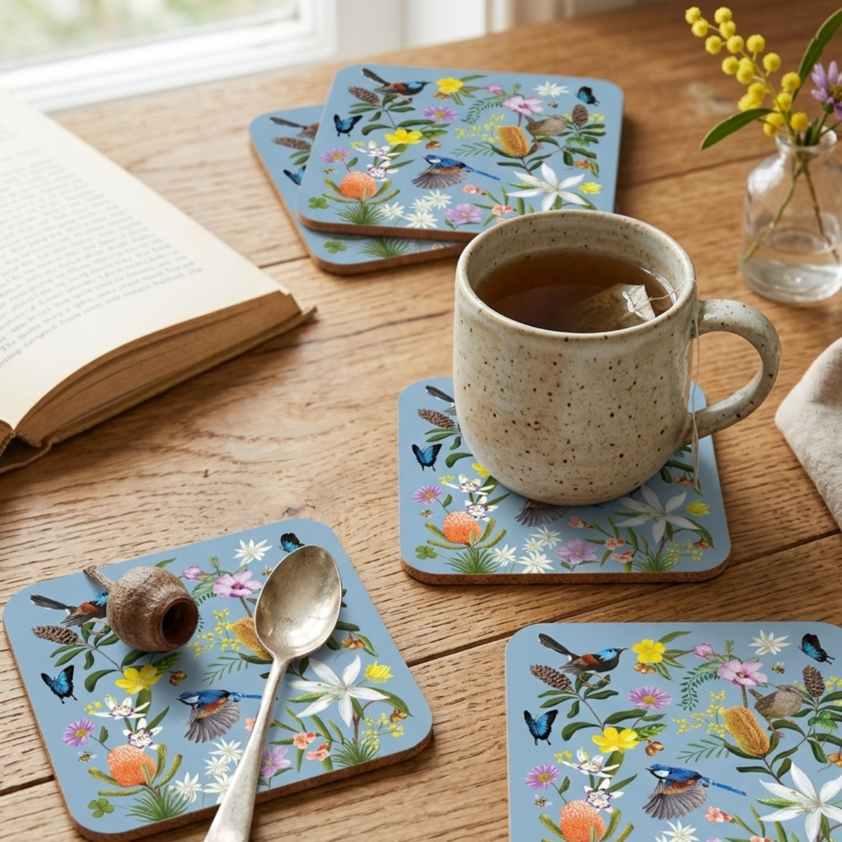 Set of floral coasters on a wooden table with a cup of tea and book.