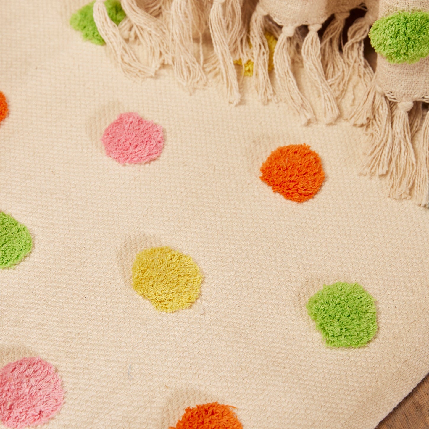 A close-up of a cream-colored woven rug with colorful tufted polka dots in pink, orange, yellow, and green, featuring a matching fringed textile draped over the top edge.