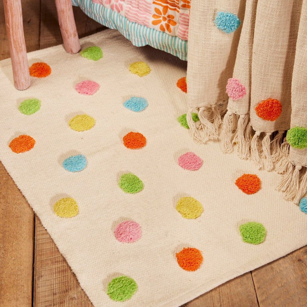 A cream-colored area rug with colorful pom-pom polka dots in green, orange, pink, yellow, and blue, styled on a wooden floor next to a wooden stool and a beige throw blanket.