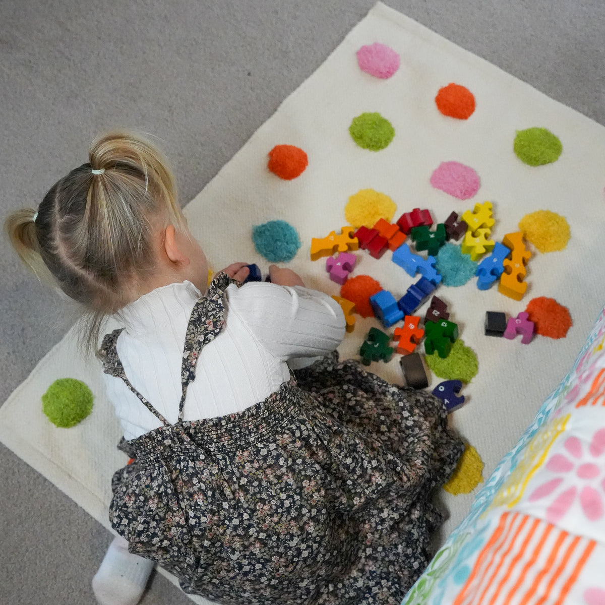 A young child with blonde hair in a ponytail sits on a cream-colored felt rug, playing with colorful toy blocks. The rug features textured felt patches in pink, green, orange, and yellow.
