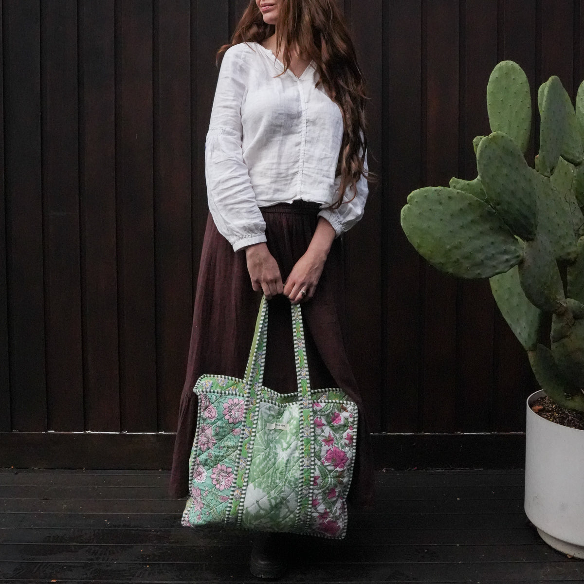 A woman wearing a white blouse and a long brown skirt holds a large green tote bag with pink floral embroidery and white beaded handles, standing in front of a dark wooden wall next to a potted cactus.