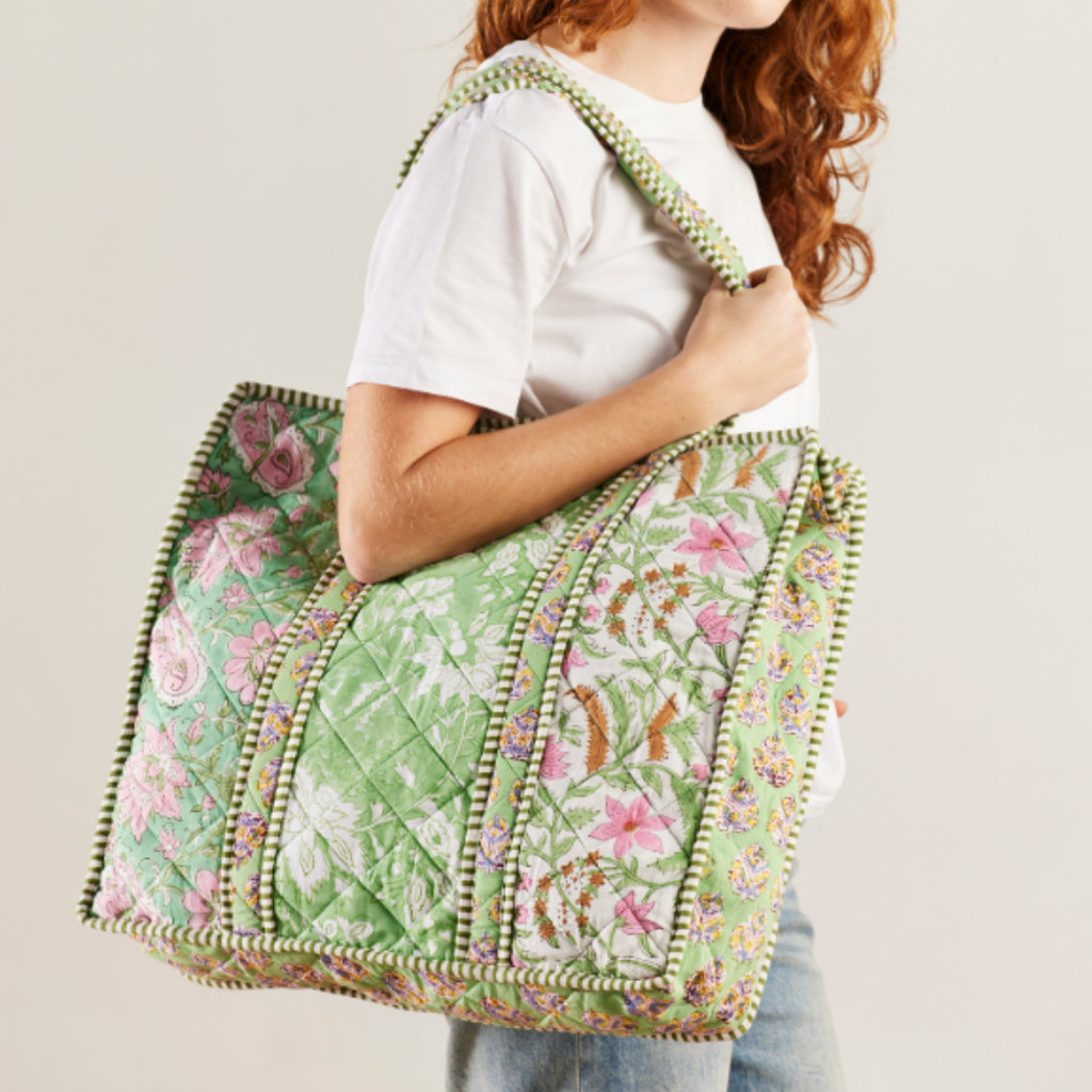 A person wearing a white t-shirt and blue jeans carries a large quilted green tote bag with pink floral patterns and a green pom-pom trim against a light beige background.