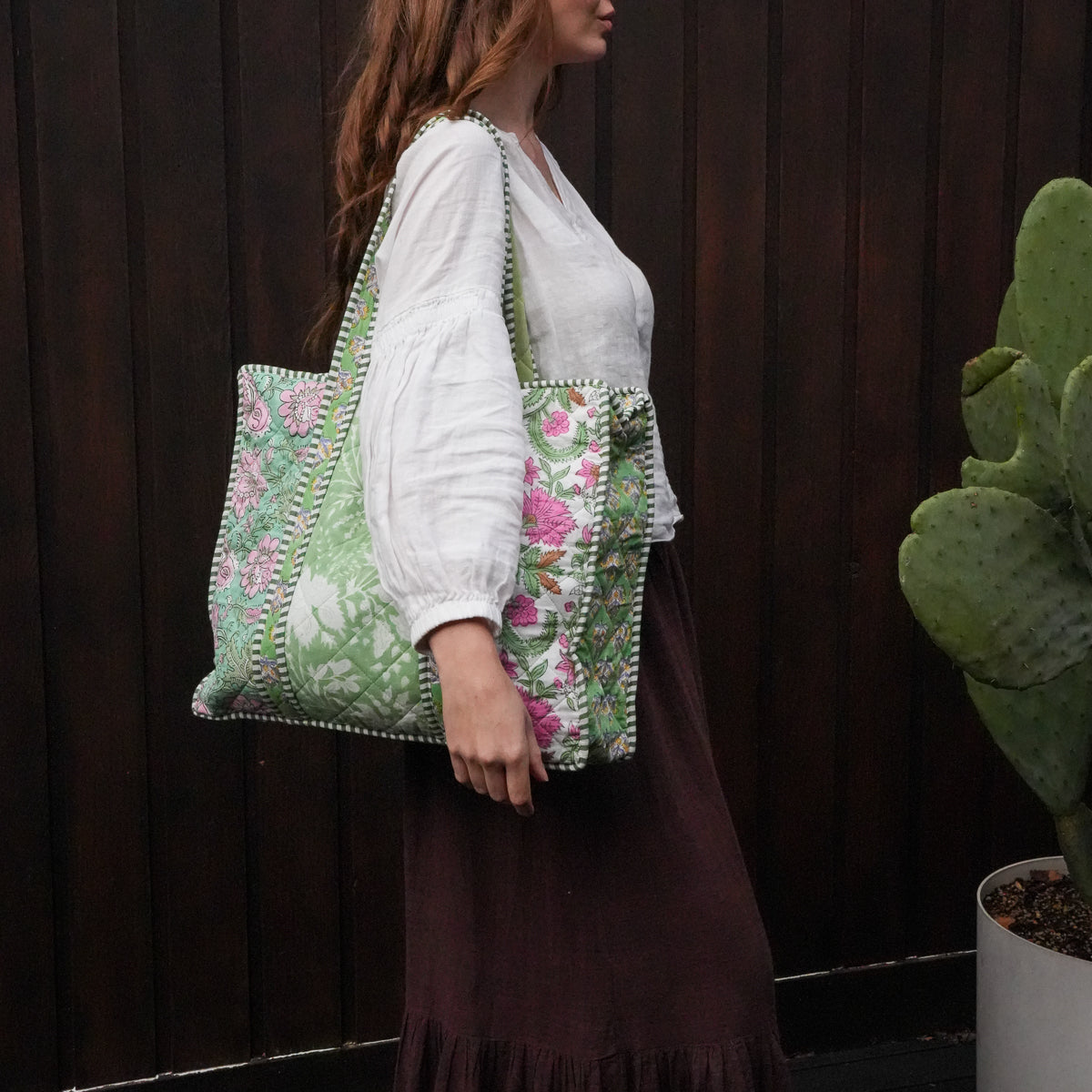 A woman wearing a white blouse and dark brown skirt carries a large green and pink floral embroidered tote bag with white pom-pom trim, standing in front of a dark wooden fence next to a potted cactus.