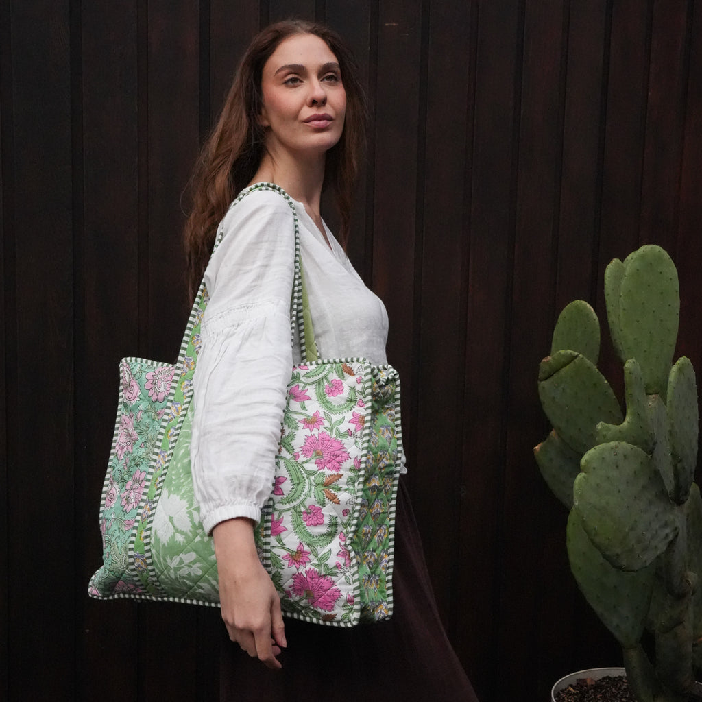 A woman wearing a white blouse and dark skirt carries a large embroidered tote bag with green floral patterns and pom-pom trim, standing against a dark wooden wall next to a potted cactus.
