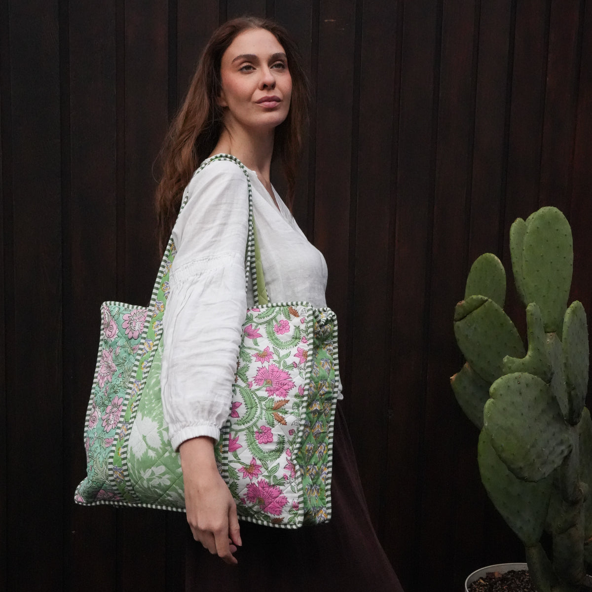A woman wearing a white blouse and dark skirt carries a large embroidered tote bag with green floral patterns and pom-pom trim, standing against a dark wooden wall next to a potted cactus.
