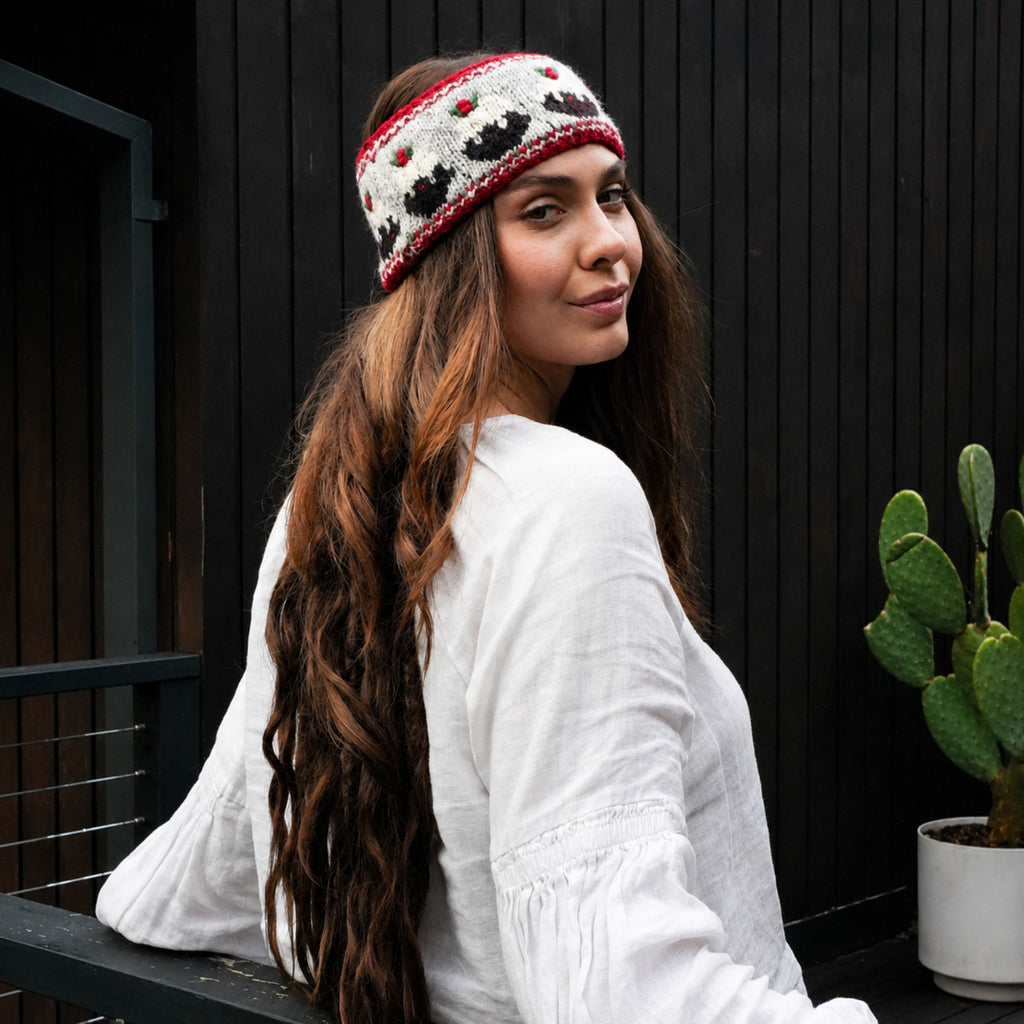 A woman wearing a white knit beanie with red and black patterns, posing outdoors against a dark wooden wall next to a potted cactus.