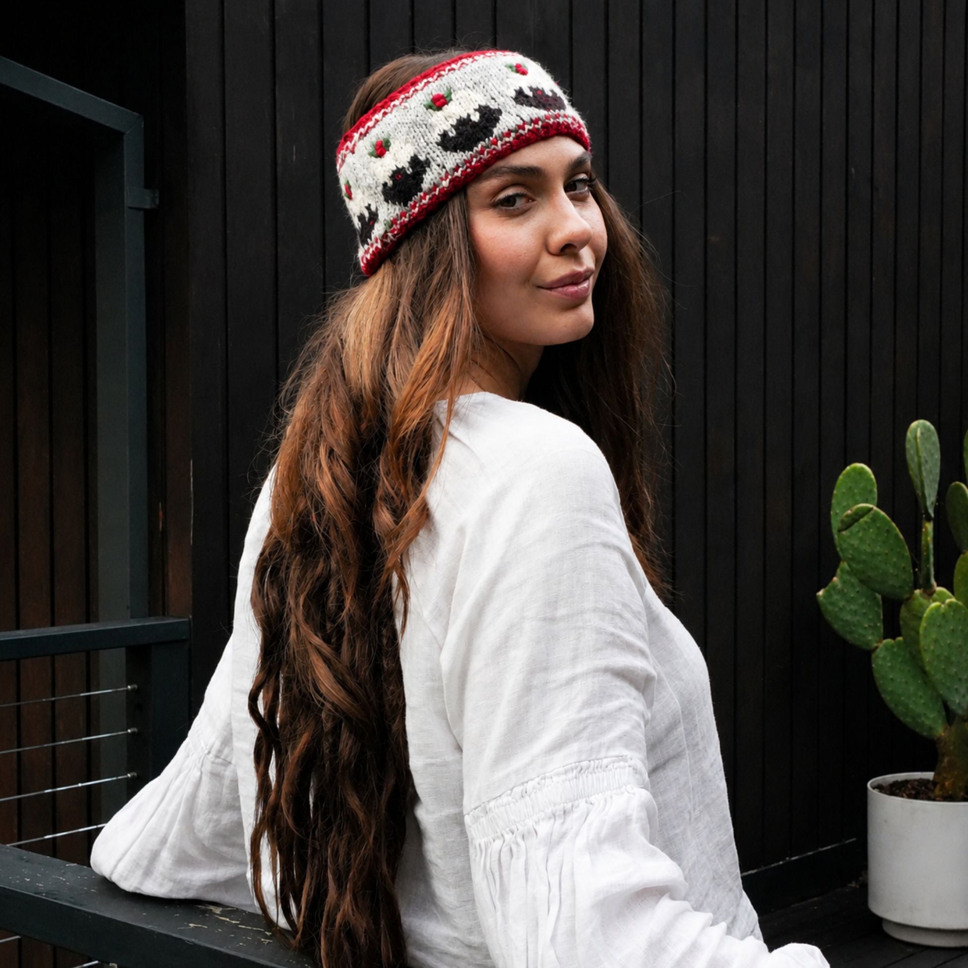 A woman wearing a white knit beanie with red and black patterns, posing outdoors against a dark wooden wall next to a potted cactus.