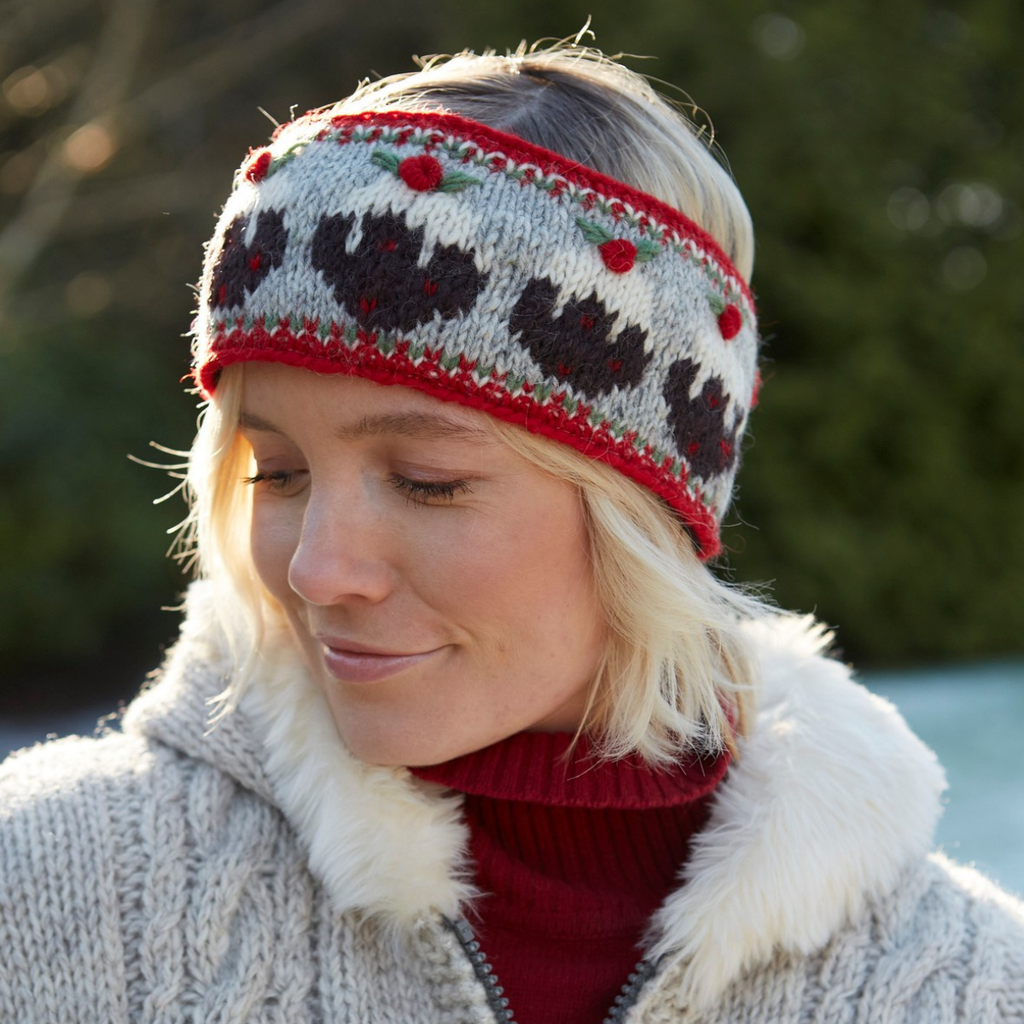 A woman wearing a knit winter headband with a red, gray, and black pattern, styled with a cream faux-fur trim and a red turtleneck under a chunky knit cardigan.