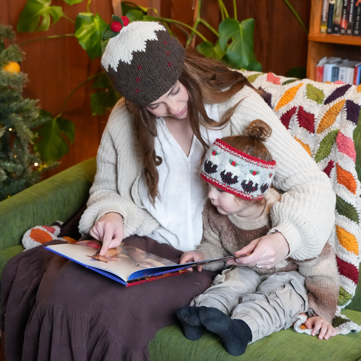 A woman and a young child sitting together on a green couch, reading a book. Both are wearing matching knit winter hats with a brown and white design and small red accents.