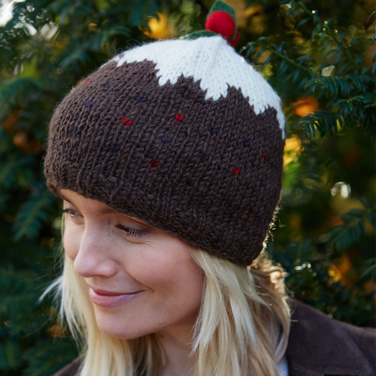 A woman wearing a knitted Christmas pudding hat with a dark brown body, white frosting-like top, and small red berries, photographed outdoors in front of green foliage.
