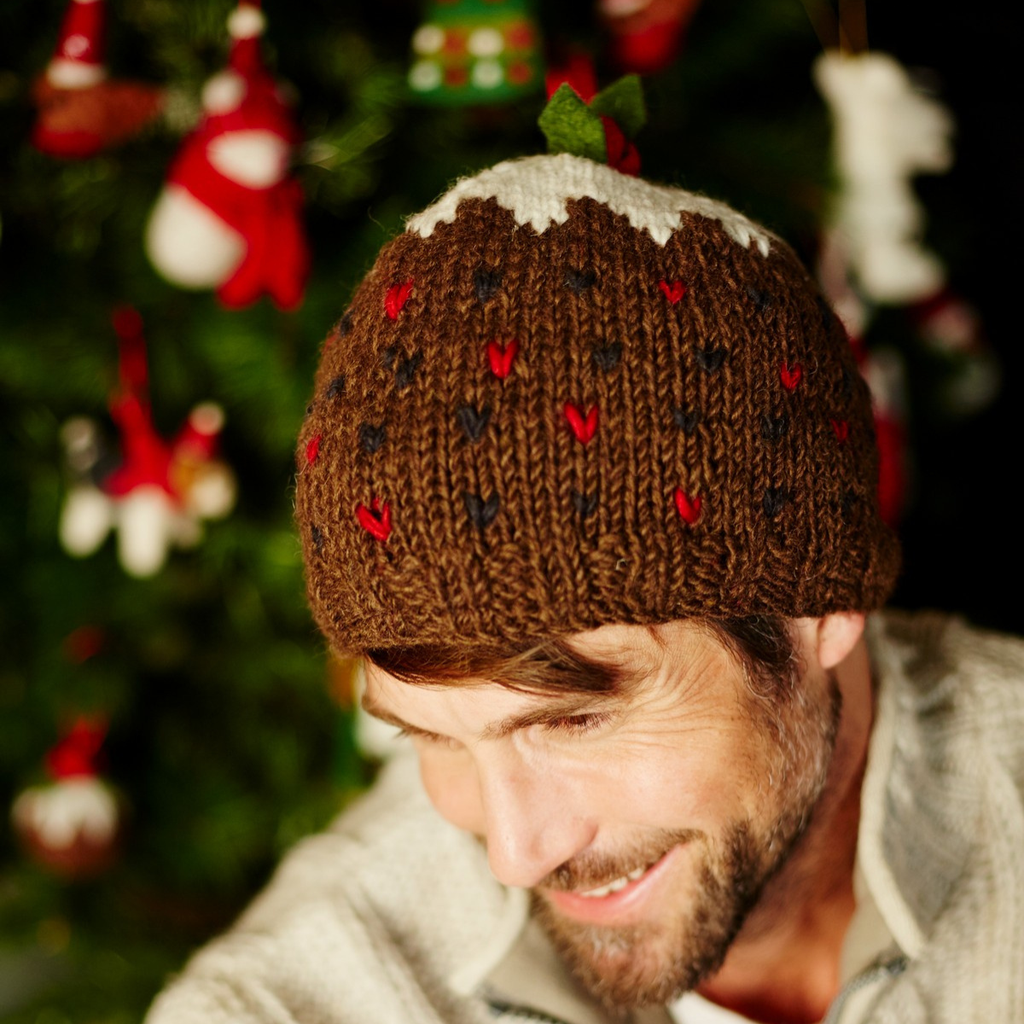 A man wearing a brown knit beanie with a white snow-like top and small red and black heart patterns, posed in front of a blurred Christmas tree with ornaments.