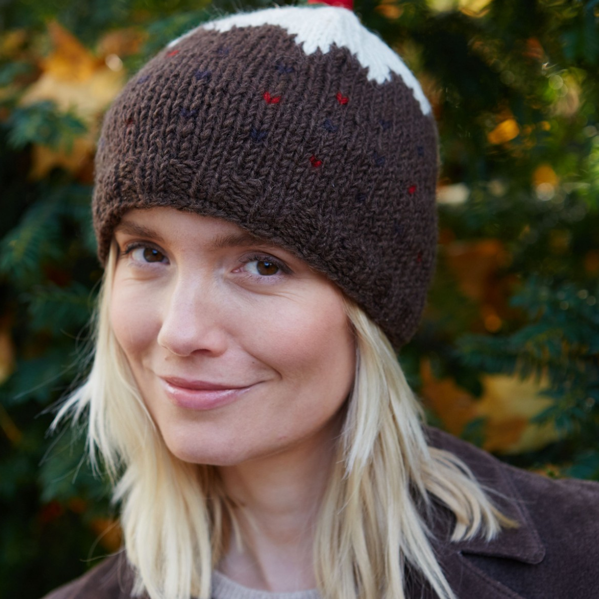 A woman wearing a brown knit beanie with a white mountain peak and small multicolored speckles, photographed outdoors with blurred greenery and autumn leaves in the background.