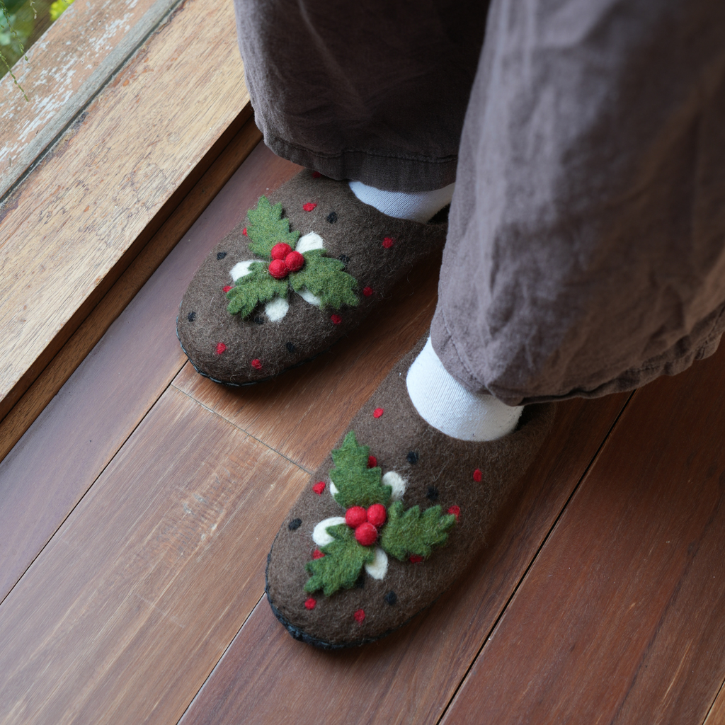 A person wearing brown felt slippers with a holly and berry pattern, standing on a wooden floor.