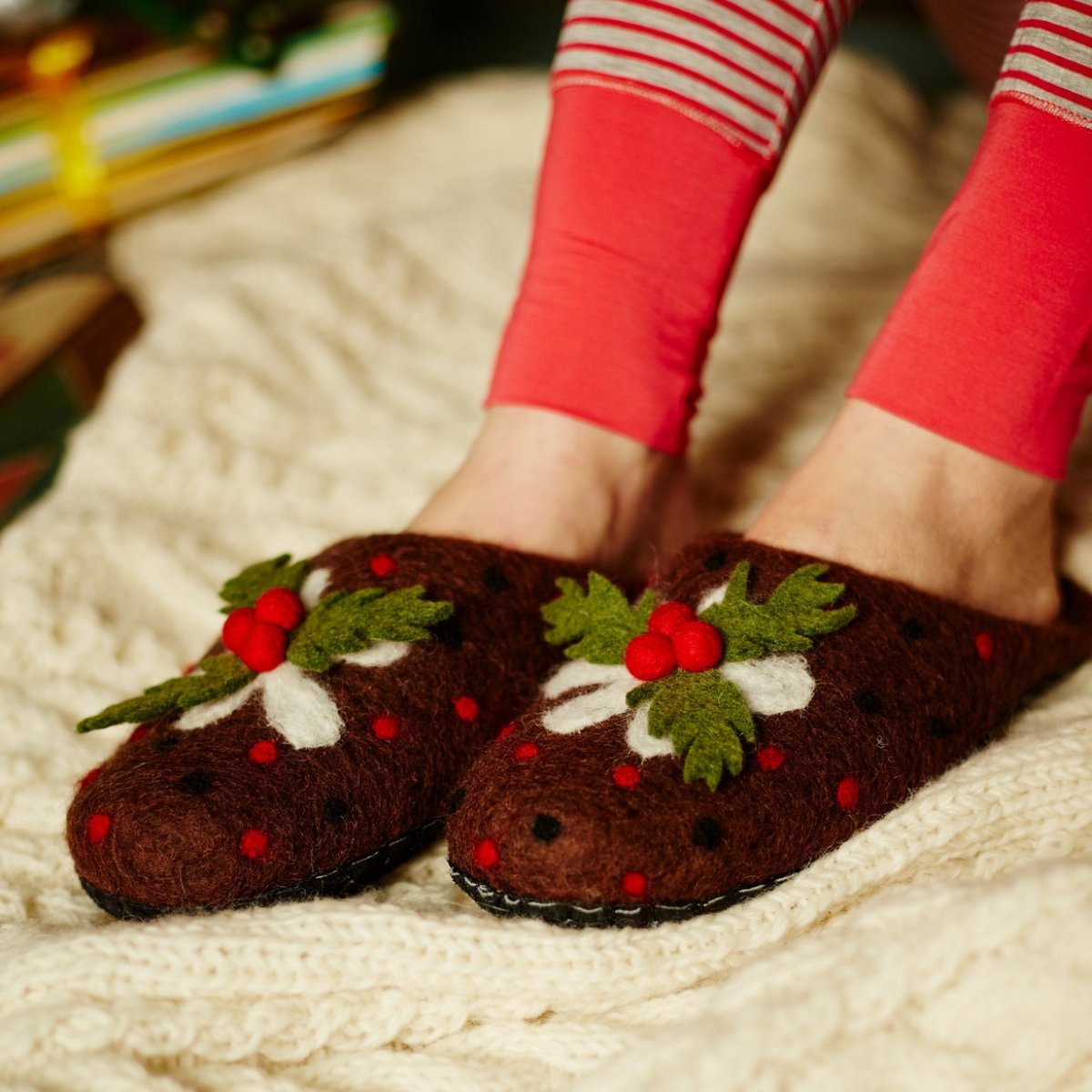 A person wearing brown felted slippers decorated with green holly leaves, white accents, and red berries, sitting on a cream cable-knit blanket.